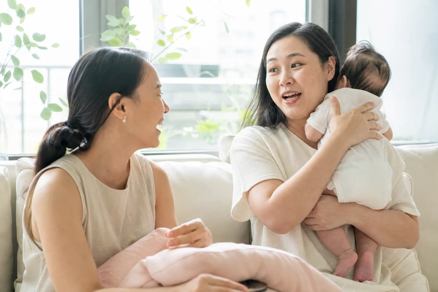 A mother holds her baby against her shoulder after a feed, turning mid-sentence towards a friend sitting beside her on a bright sofa.