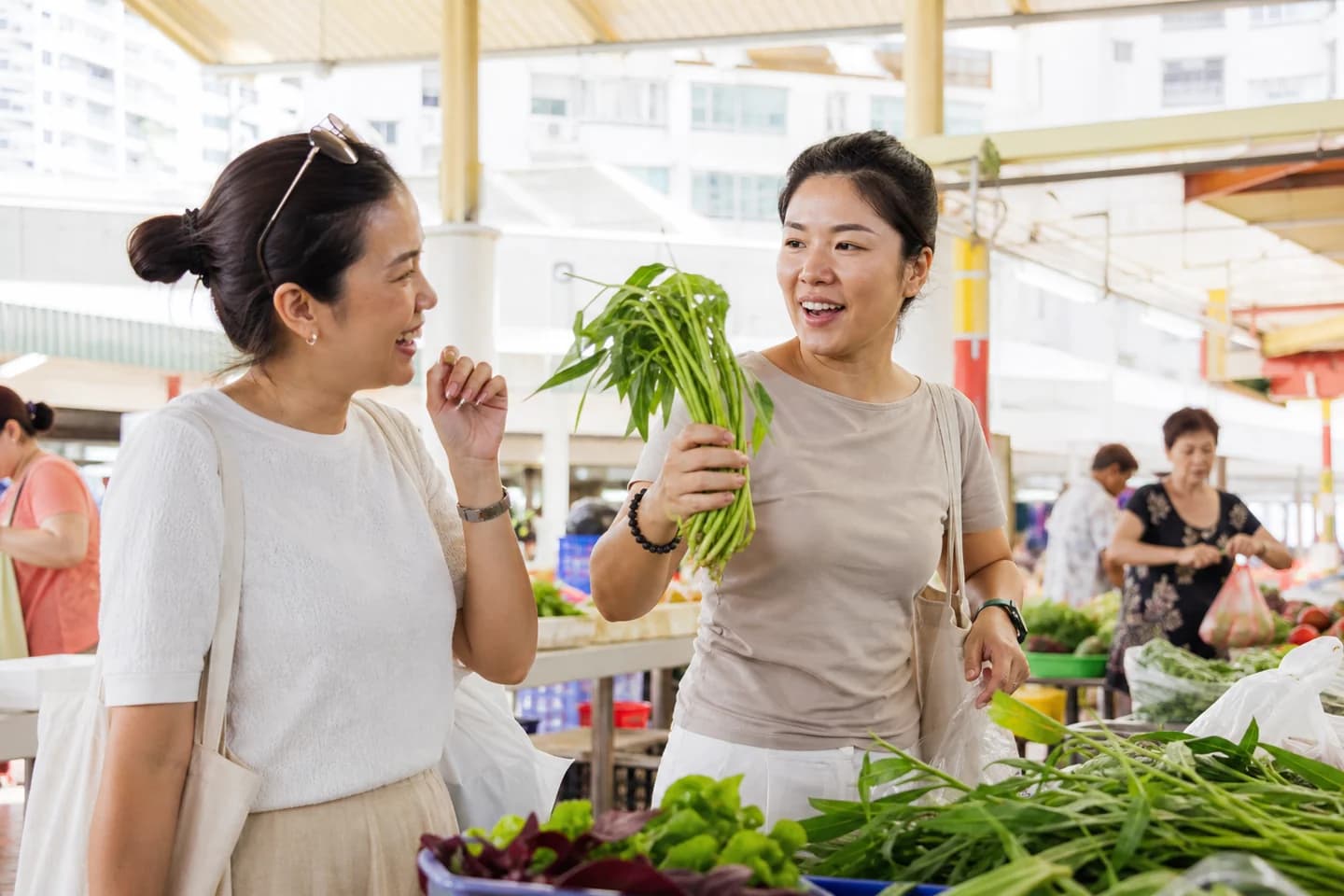Two women choose vegetables together at a covered wet market in the morning, one holding up a bunch of greens mid-sentence while the other laughs.