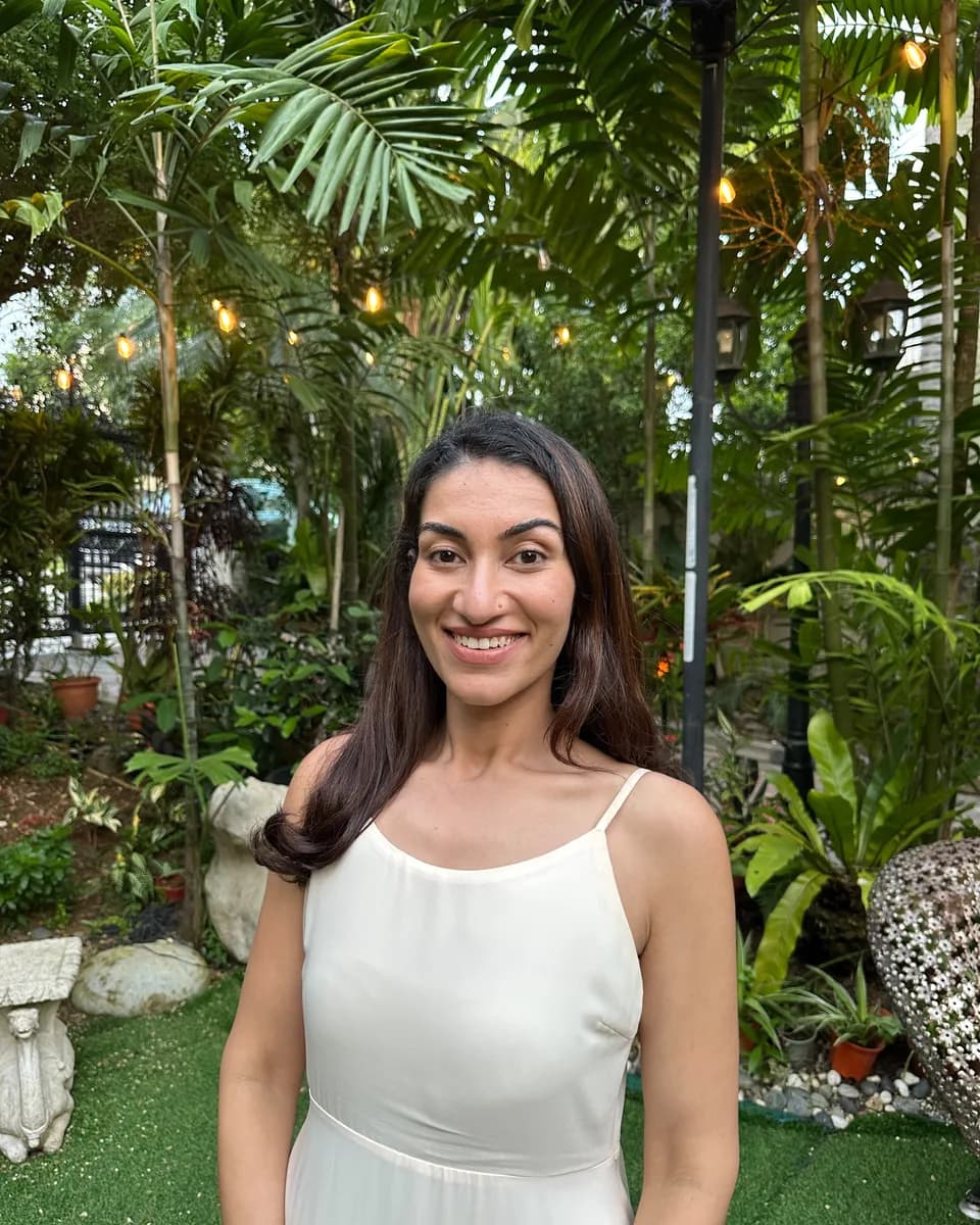 Dr Kavina Shahi smiling at the camera in a cream dress, in a garden at dusk with palms and string lights behind her.