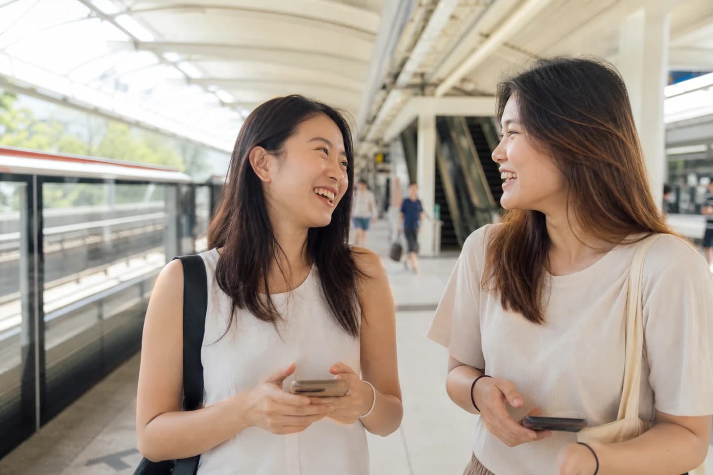 Two young women stand together on a bright open MRT platform mid-morning, one turning to the other and laughing.