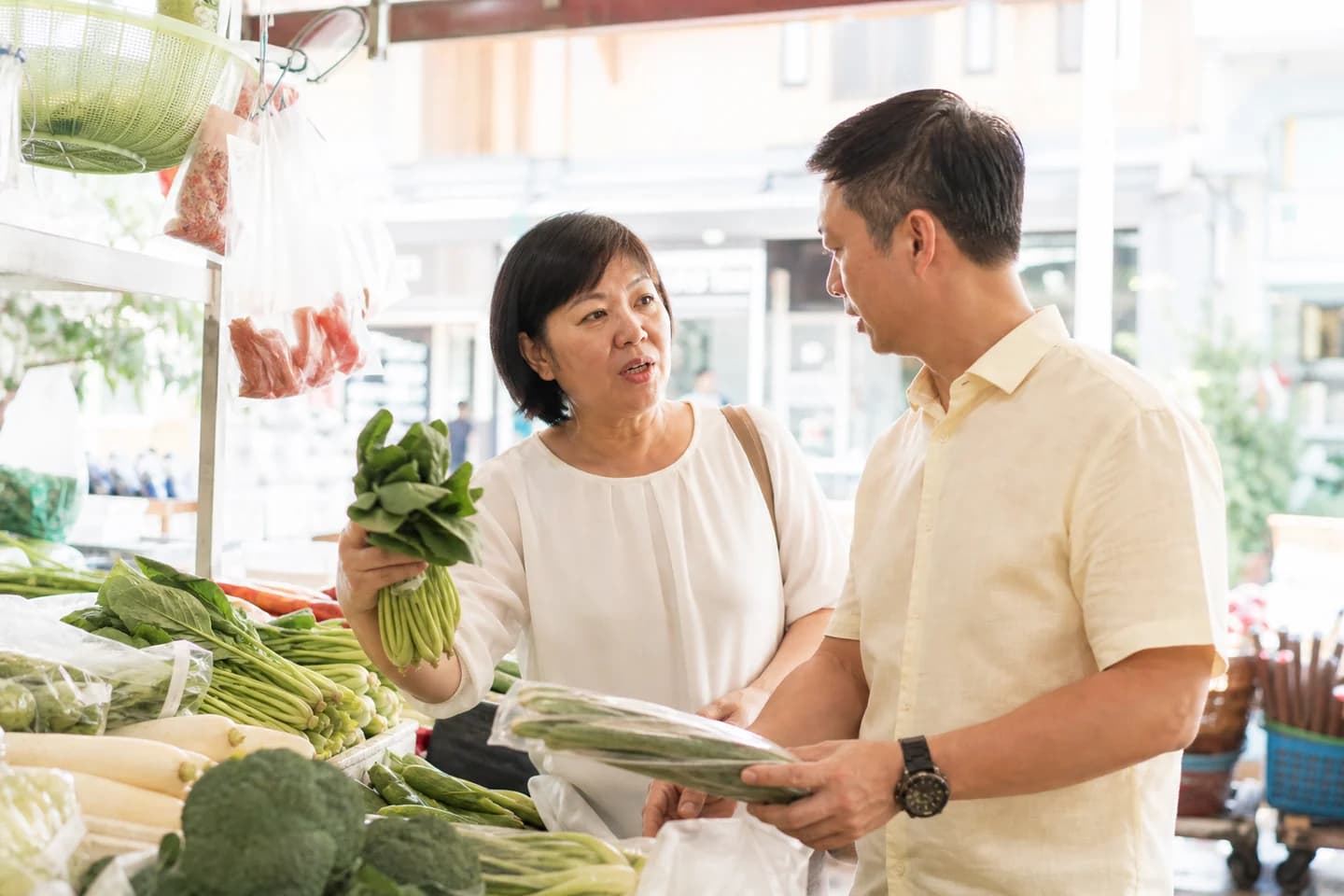 A woman in her forties and her husband choose vegetables together at a bright greengrocer, both holding produce, mid-conversation.