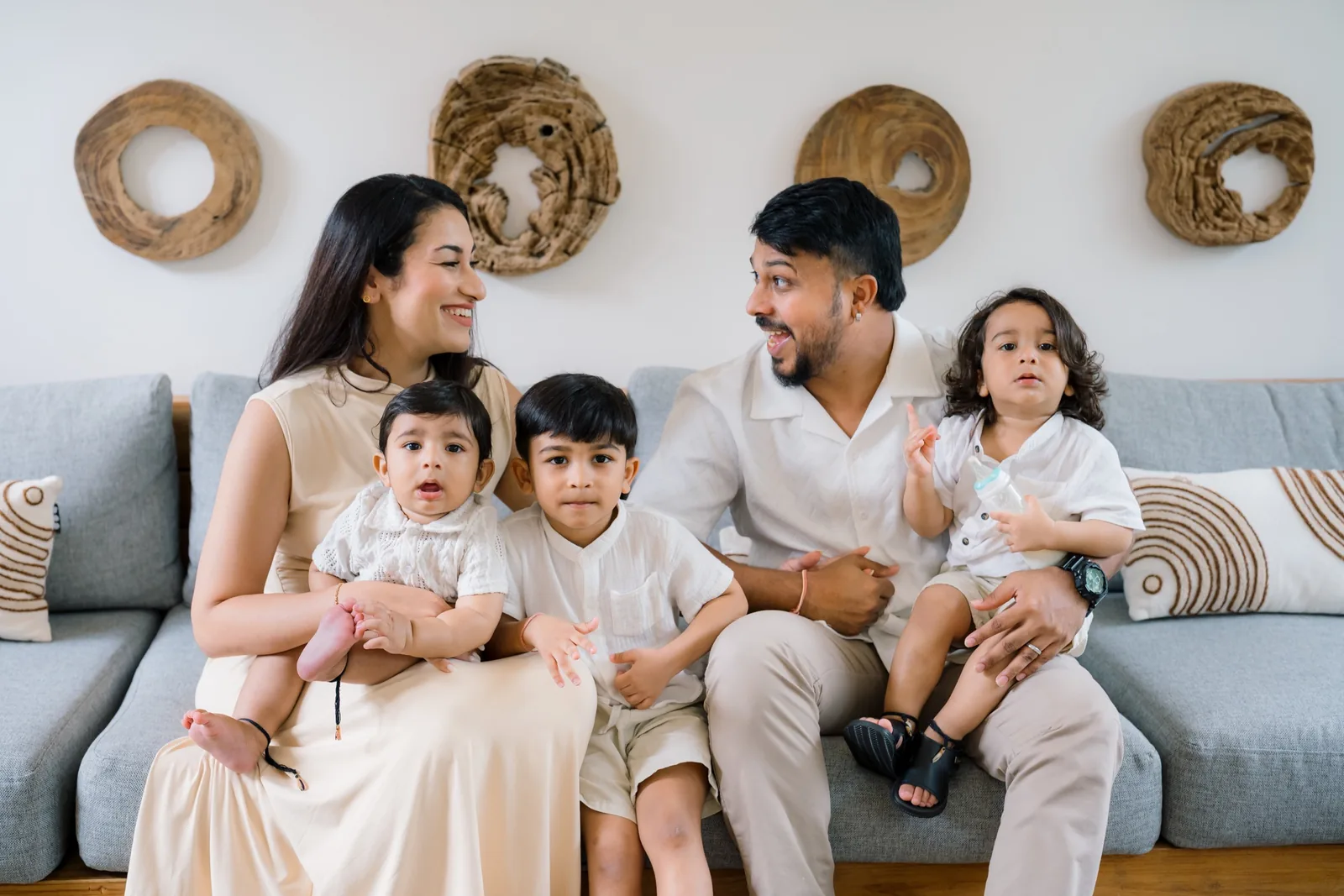 Dr Kavina Shahi with her husband and three young sons on a grey sofa at home, everyone in cream and white, laughing together.