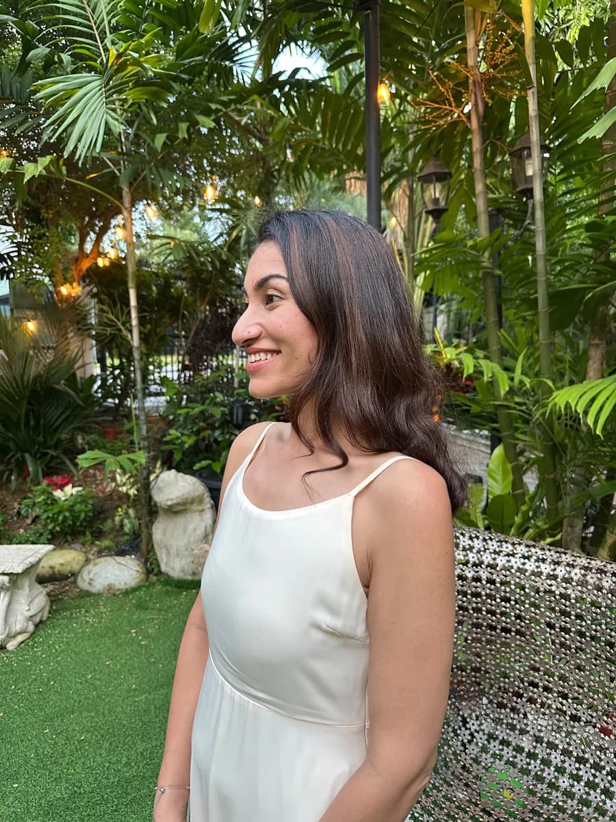 Dr Kavina Shahi in profile in a tropical garden at golden hour, smiling and looking away from the camera, in an ivory dress with string lights in the palms behind her.