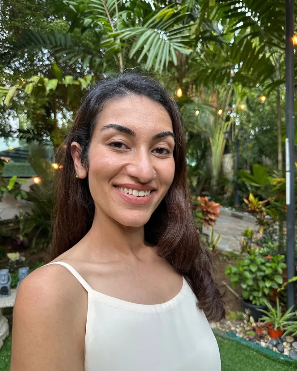 Dr Kavina Shahi smiling at the camera in a tropical garden at golden hour, in an ivory dress, with string lights and palms behind her.