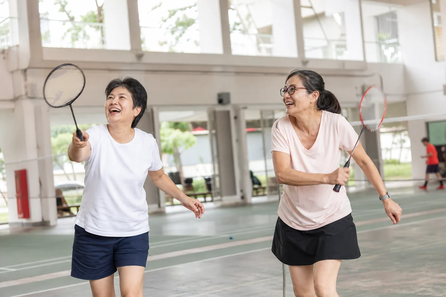Two women in their fifties mid-rally on a bright community centre badminton court, rackets up, both laughing.