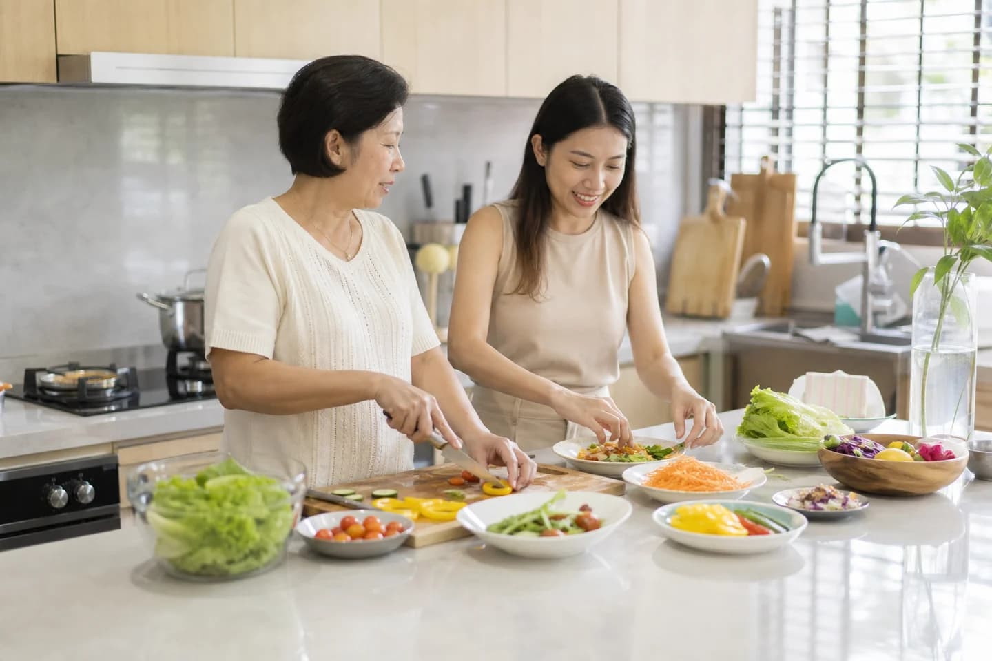 A woman in her fifties and her adult daughter prepare a colourful meal together at a bright kitchen island, both mid-action.