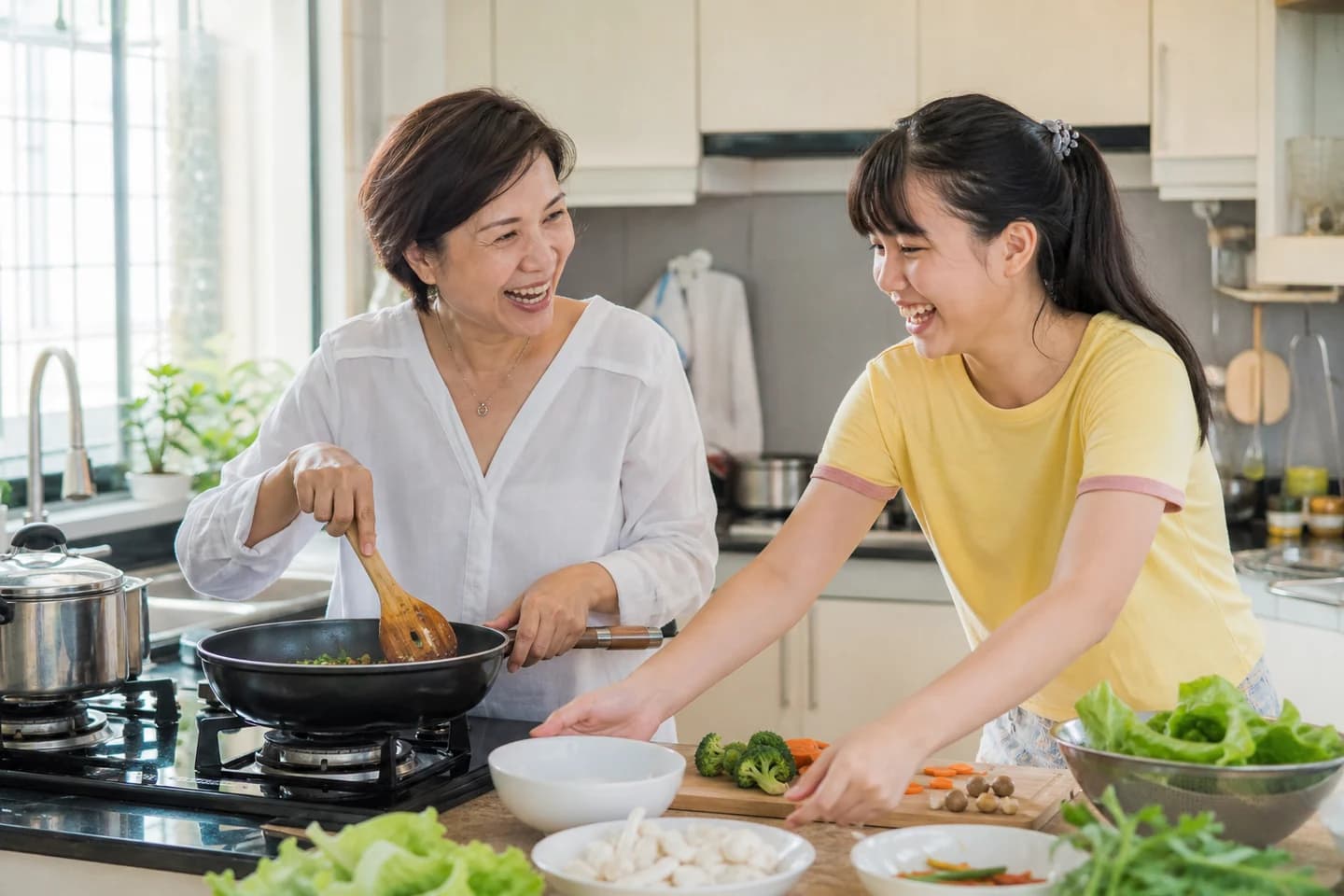 A woman in her late forties and her teenage daughter cook together in a bright kitchen, one stirring and one reaching past with a bowl.