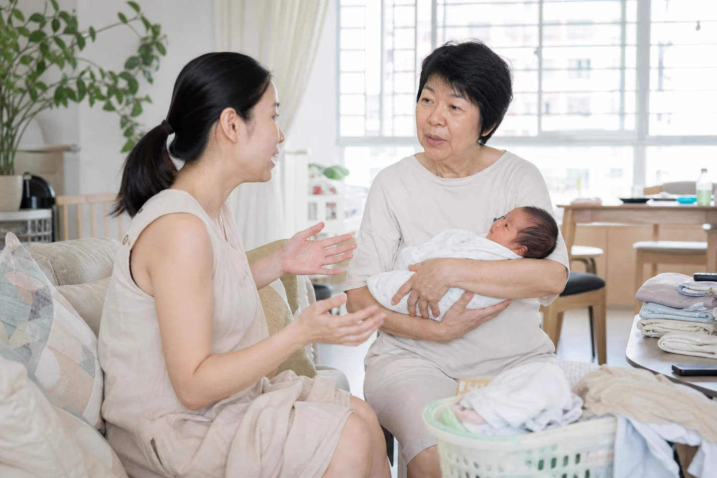 A new mother sits talking with her own mother in a bright living room at midday, the newborn in the grandmother's arms.