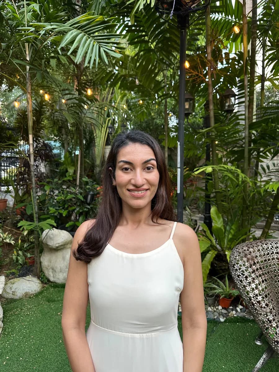 Dr Kavina Shahi in a cream dress smiling at the camera, standing on a lawn in a garden at dusk, palms and string lights behind her.