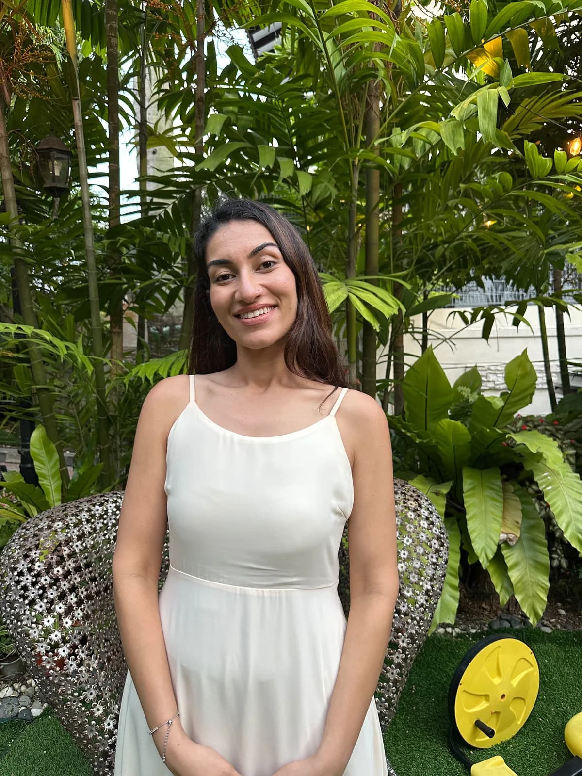 Dr Kavina Shahi standing in a tropical garden at golden hour in an ivory dress, smiling at the camera, string lights in the palms behind her.